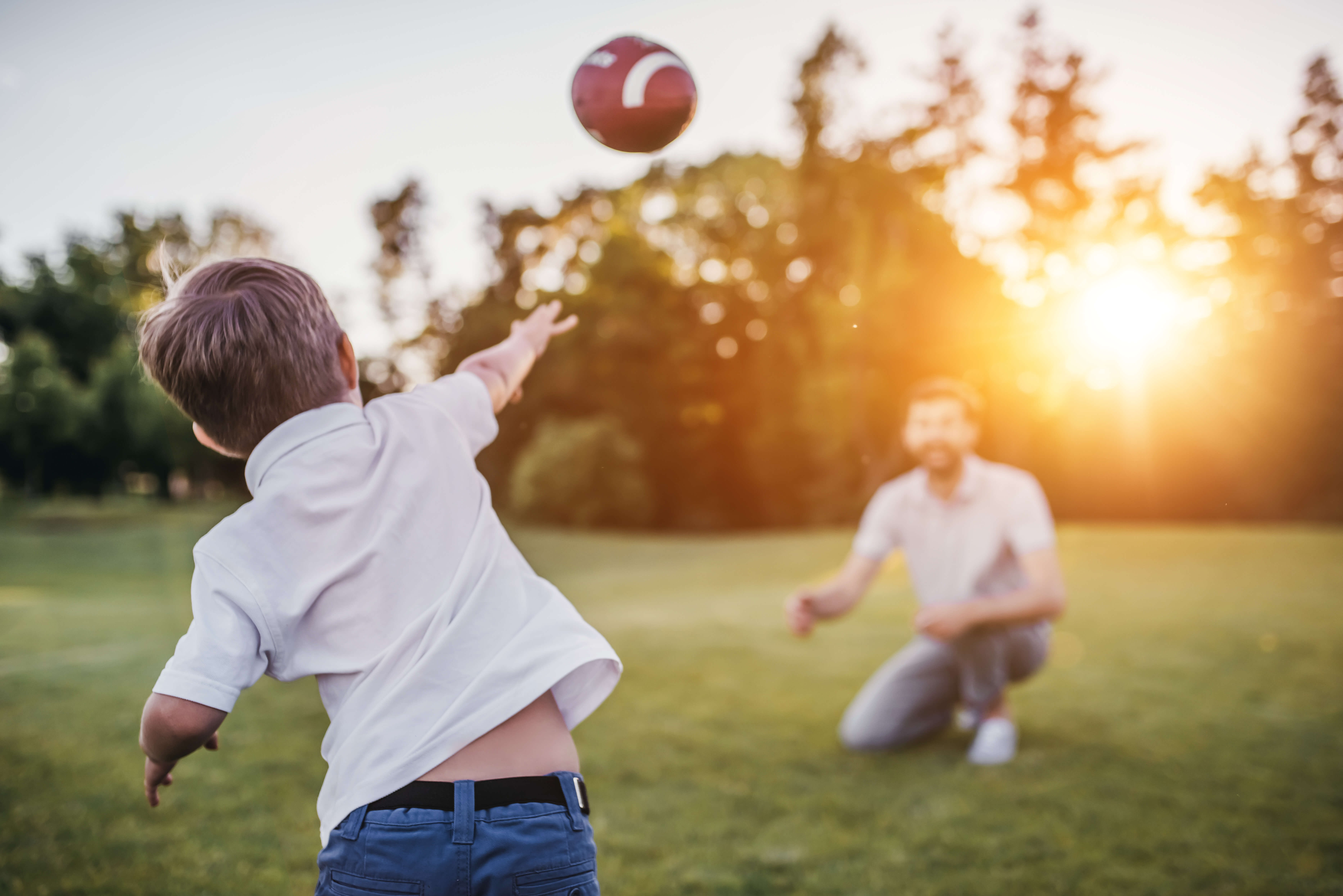 Handsome dad with his little cute sun are having fun and playing American football on green grassy lawn